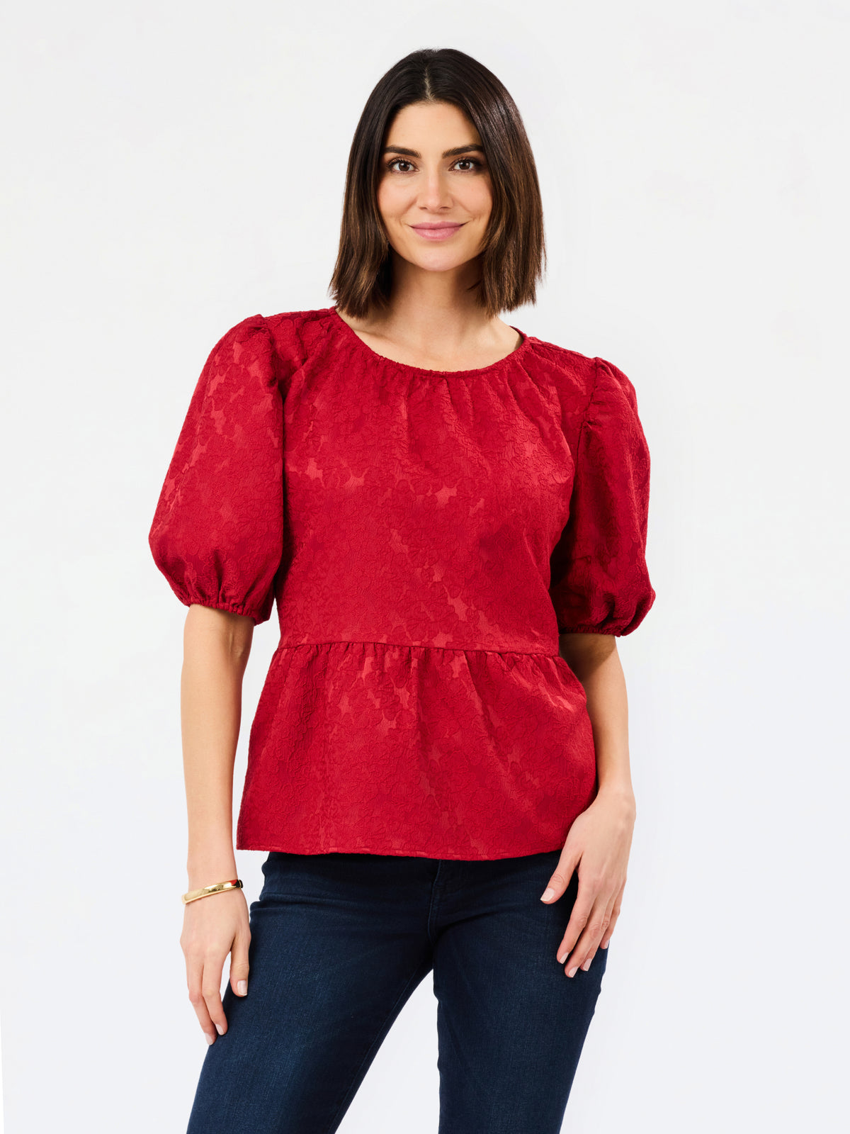 A woman modeling a red short-sleeve peplum blouse with puff sleeves and a textured, slightly mottled fabric, paired with dark blue jeans against a plain white background.