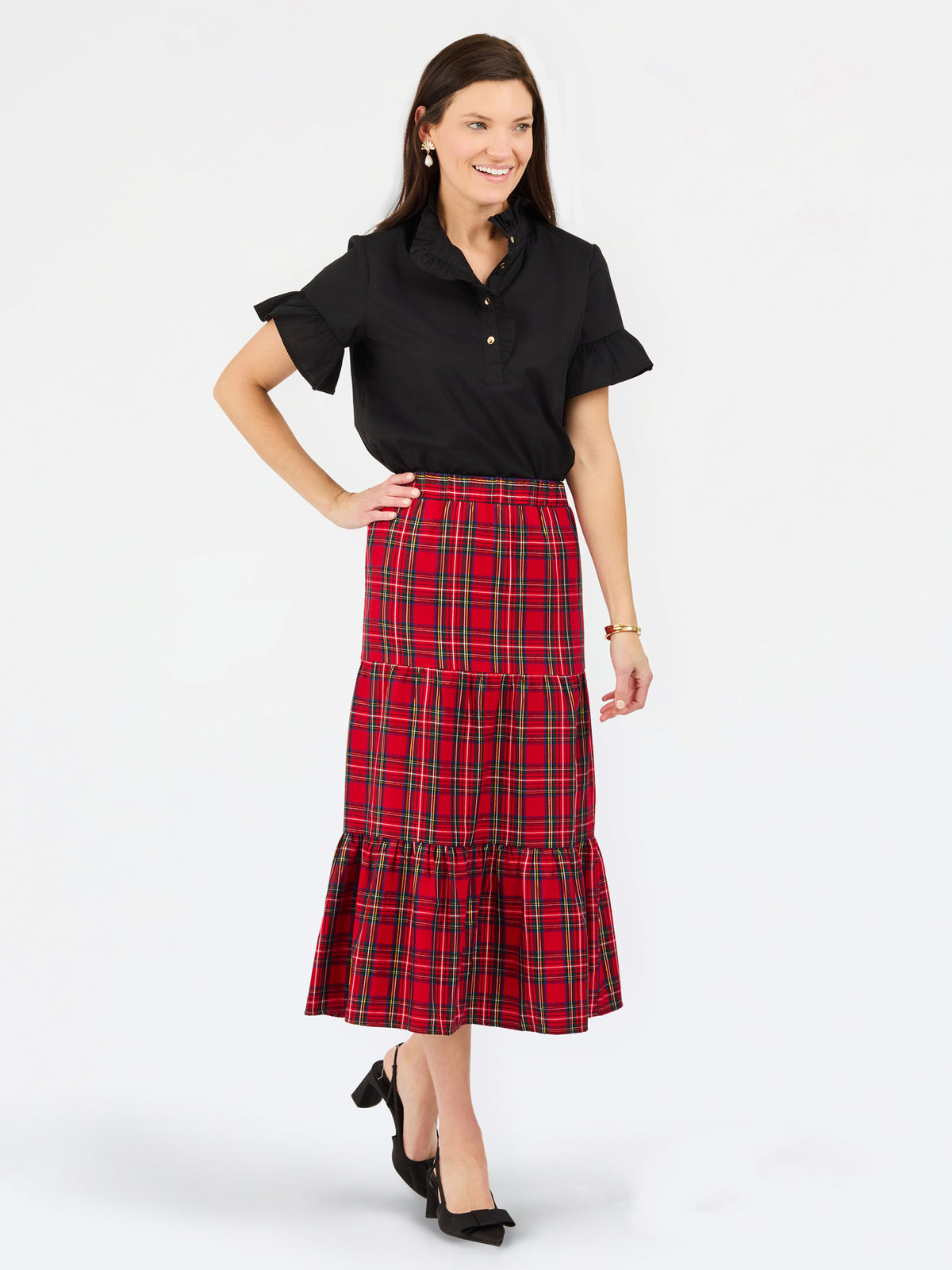 A woman modeling a black short-sleeve collared shirt and a red tiered plaid midi skirt with black heels, photographed against a light gray studio background.
