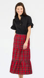 A woman modeling a black short-sleeve button-up blouse with ruffled sleeves and a red and black plaid tiered midi skirt, photographed against a plain light gray studio background.
