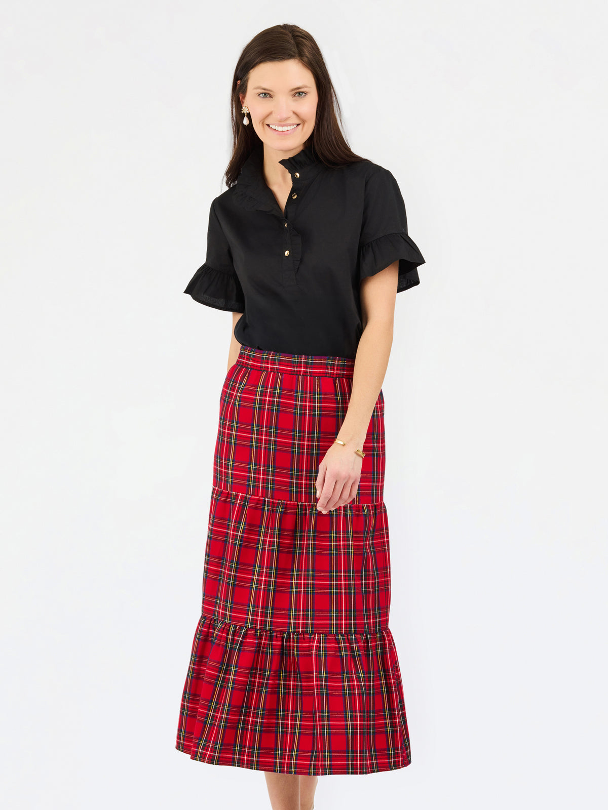 A woman modeling a black short-sleeve button-up blouse with ruffled sleeves and a red and black plaid tiered midi skirt, photographed against a plain light gray studio background.