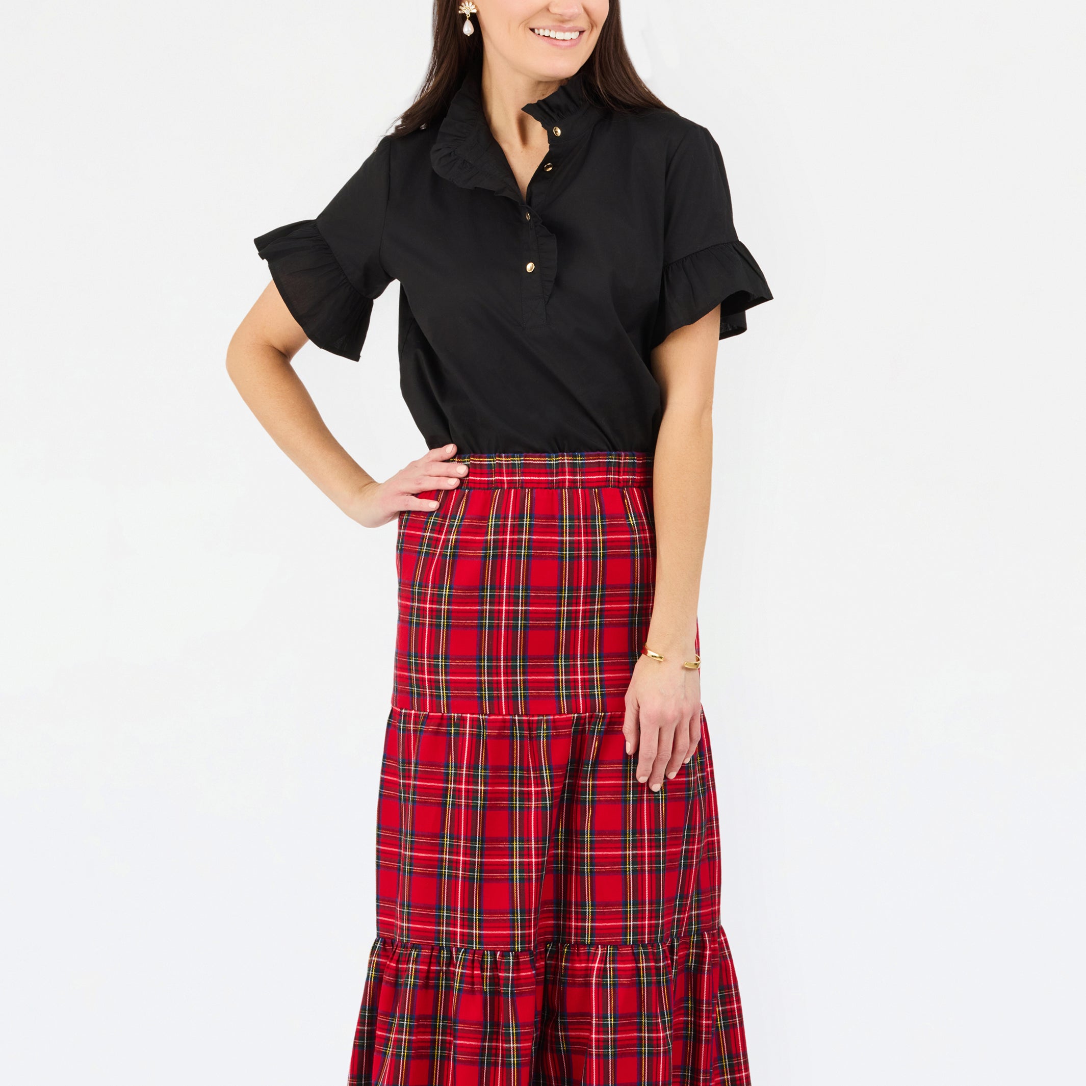 A woman modeling a black short-sleeve button-up shirt with ruffled sleeves and a high-waisted red and black plaid tiered midi skirt, set against a plain white studio background.