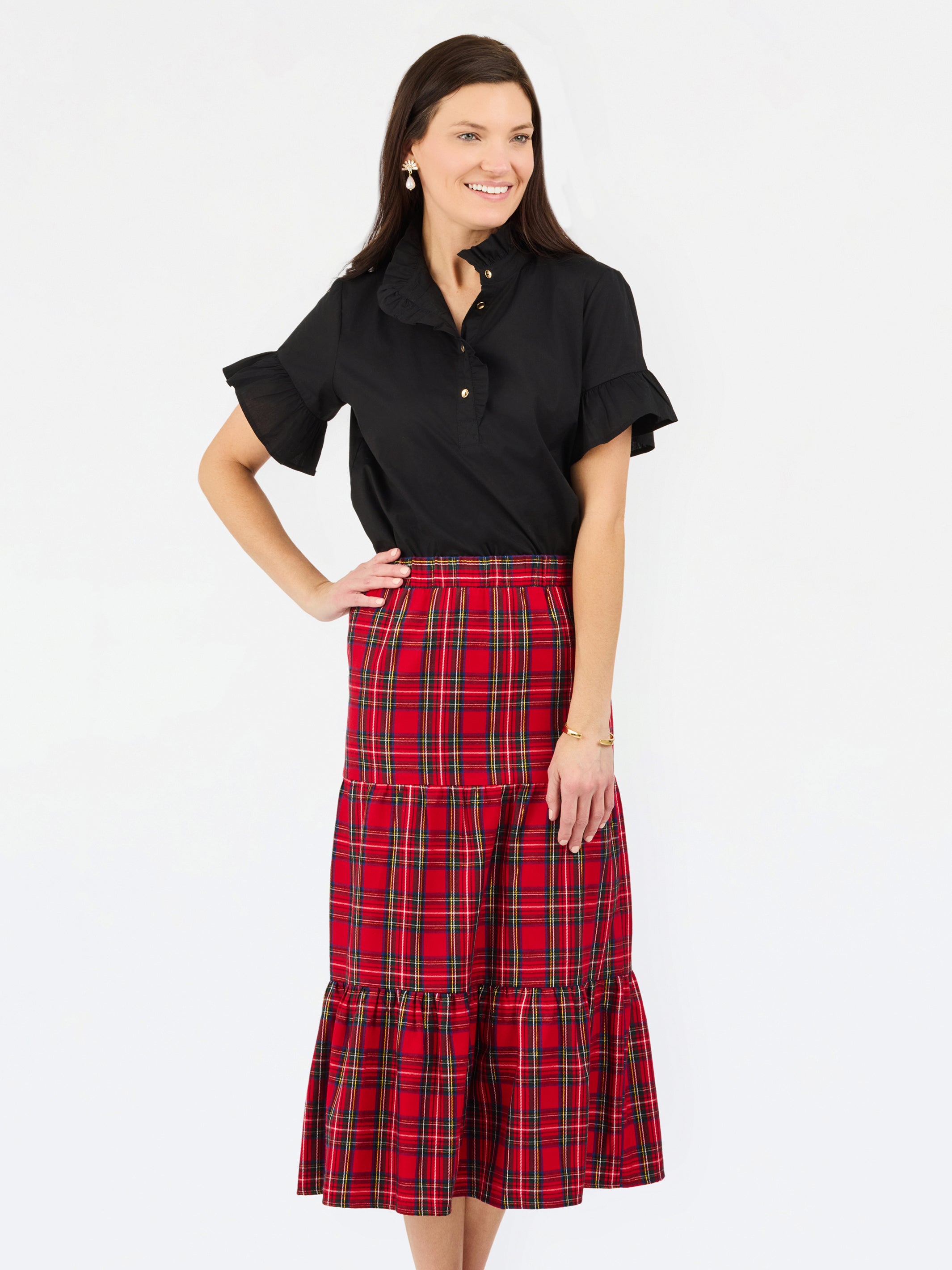 A woman modeling a black short-sleeve button-up shirt with ruffled sleeves and a high-waisted red and black plaid tiered midi skirt, set against a plain white studio background.