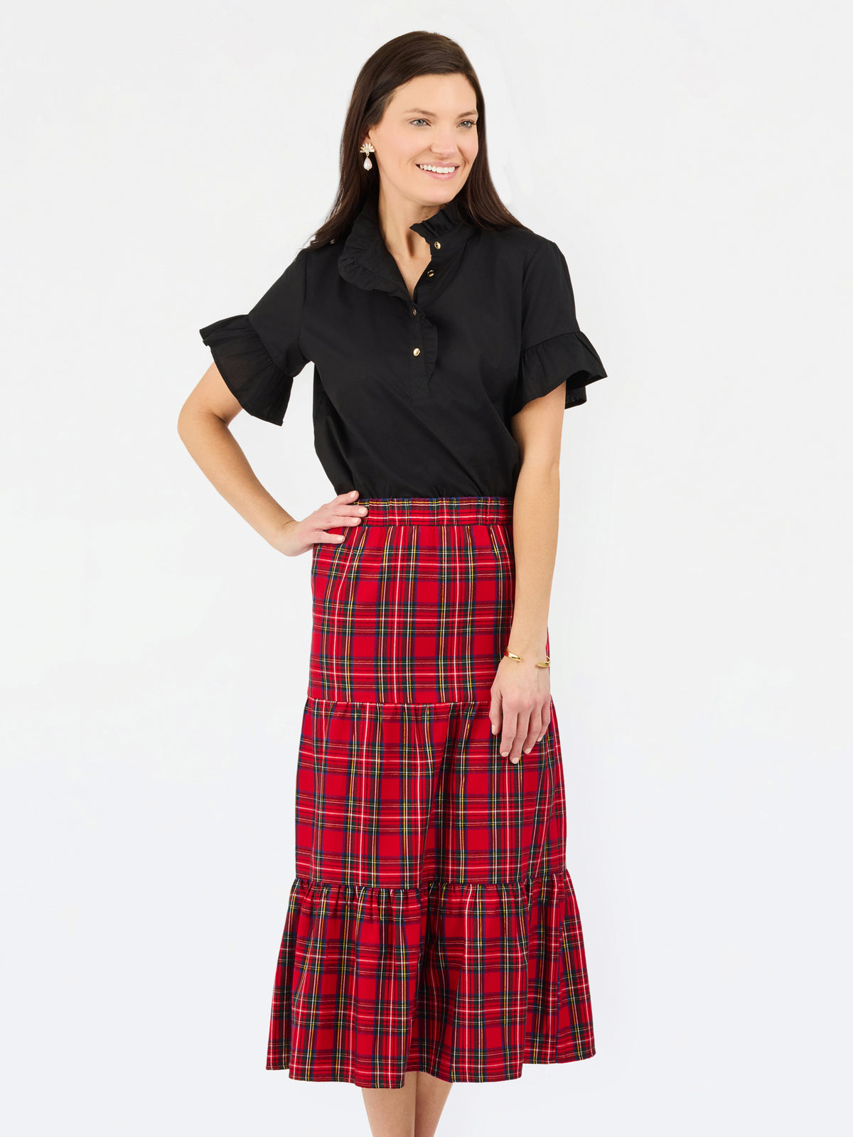 A woman modeling a black short-sleeve button-up shirt with ruffled sleeves and a high-waisted red and black plaid tiered midi skirt, set against a plain white studio background.
