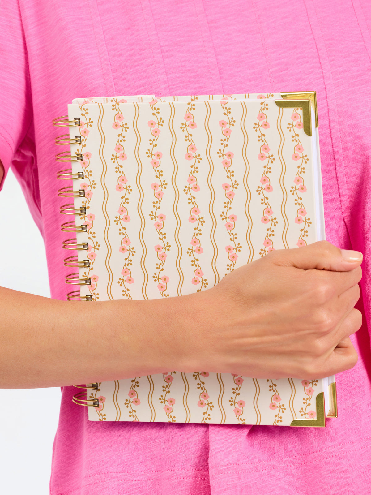 A close-up of a person in a pink top holding a spiral-bound notebook with a cream cover decorated with vertical wavy gold lines and pink floral motifs, plus gold corner accents.