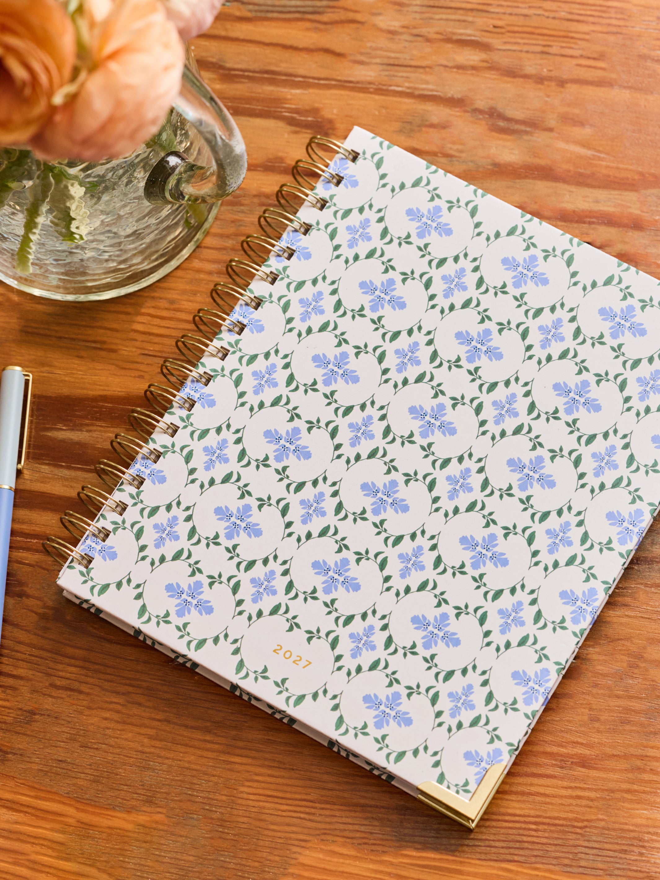 A spiral-bound planner or notebook with a white cover decorated in a repeating light blue floral pattern and green leaves, placed on a wooden tabletop next to a glass vase with flowers and part of a pen.