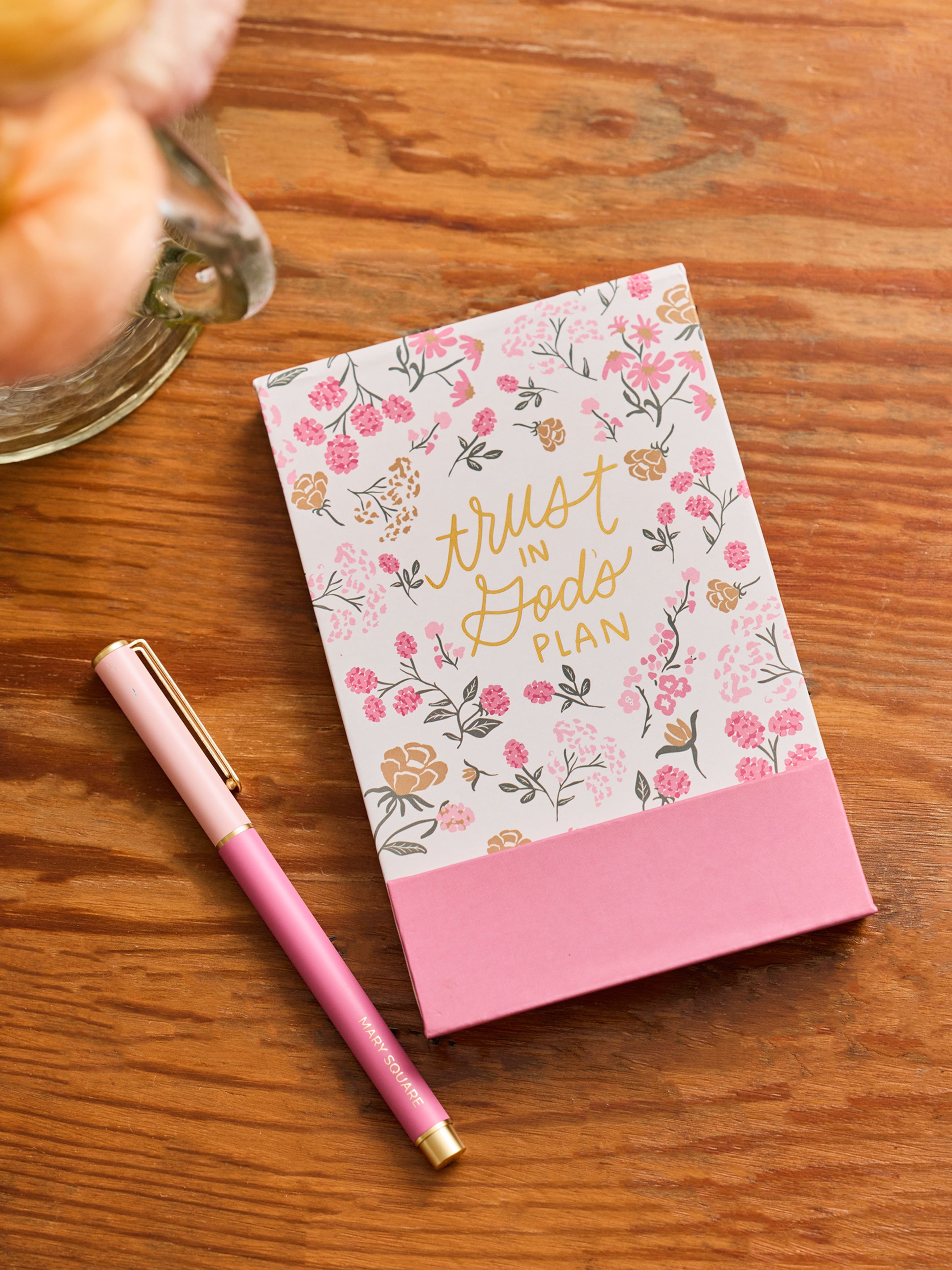A floral notepad or journal titled 'trust in God's PLAN' in gold script, placed on a wooden table next to a pink pen. The cover has a white background with pink and gold botanical illustrations, and a solid pink band at the bottom.