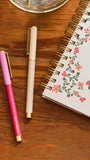 A styled flat-lay on a wooden tabletop featuring a pink and magenta pen with gold trim, a cream-colored pen with gold trim, and a spiral-bound planner with a pink and green floral pattern.