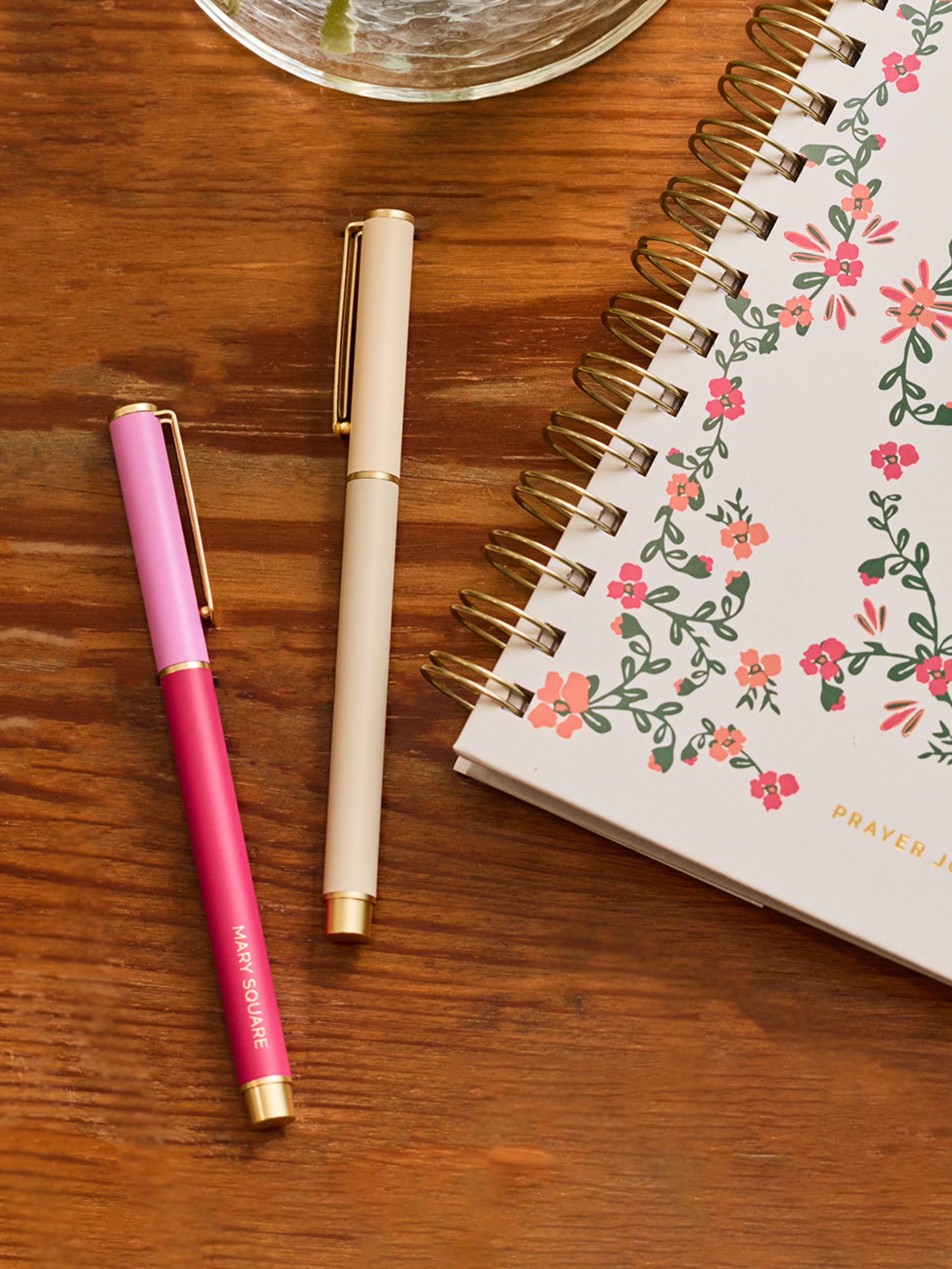 A styled flat-lay on a wooden tabletop featuring a pink and magenta pen with gold trim, a cream-colored pen with gold trim, and a spiral-bound planner with a pink and green floral pattern.