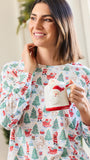 A woman wearing a festive long-sleeve pajama top with a Christmas print, holding a Santa-themed mug in a brightly lit indoor setting.