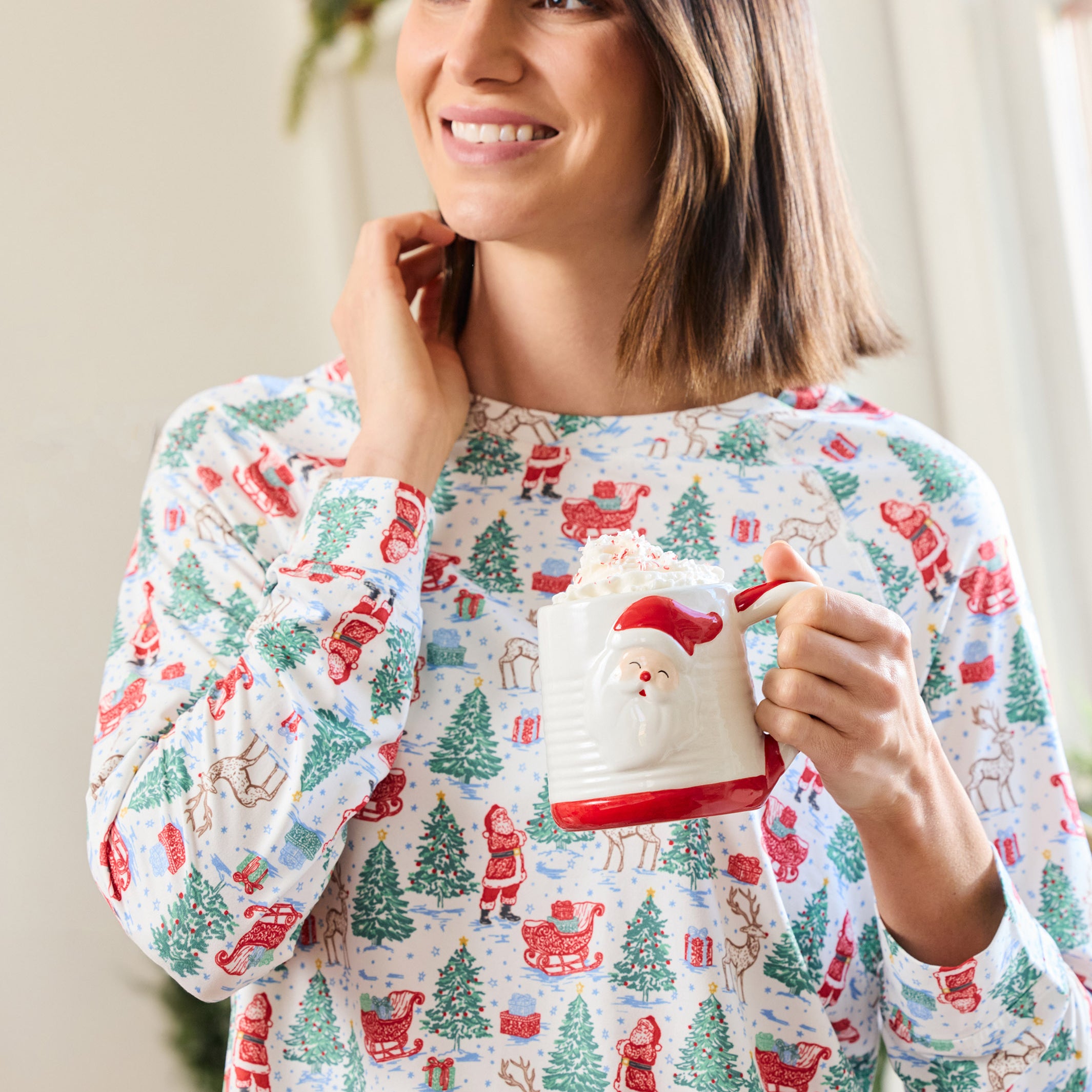 A woman wearing a festive long-sleeve pajama top with a Christmas print, holding a Santa-themed mug in a brightly lit indoor setting.