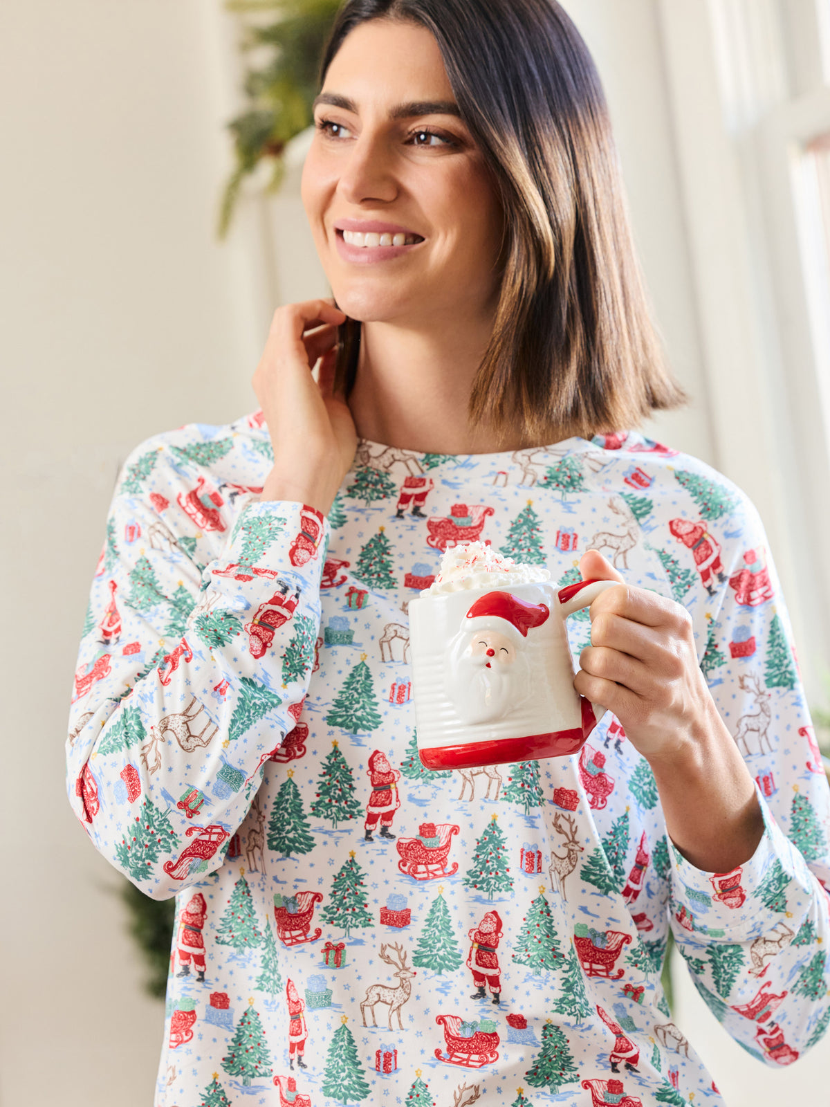 A woman wearing a festive long-sleeve pajama top with a Christmas print, holding a Santa-themed mug in a brightly lit indoor setting.