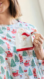 A woman in a festive holiday pajama shirt holding a white ceramic mug with a red Santa face and red handle. The pajama shirt features a repeating pattern of Santa Claus, sleighs, and green Christmas trees on a light blue background.