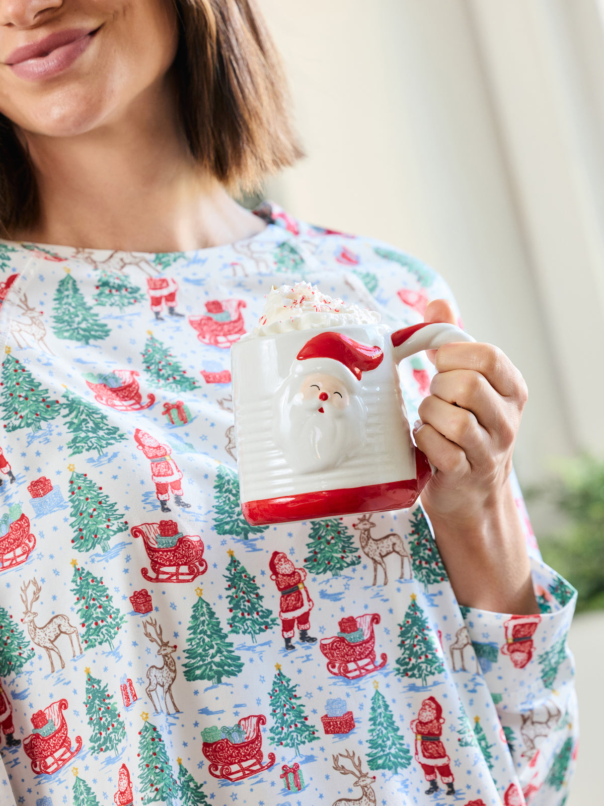 A woman in a festive holiday pajama shirt holding a white ceramic mug with a red Santa face and red handle. The pajama shirt features a repeating pattern of Santa Claus, sleighs, and green Christmas trees on a light blue background.