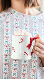 A person wearing a festive holiday sweater with a red, green, and white candy cane and bow pattern, holding a white mug filled with whipped cream and a red handle.