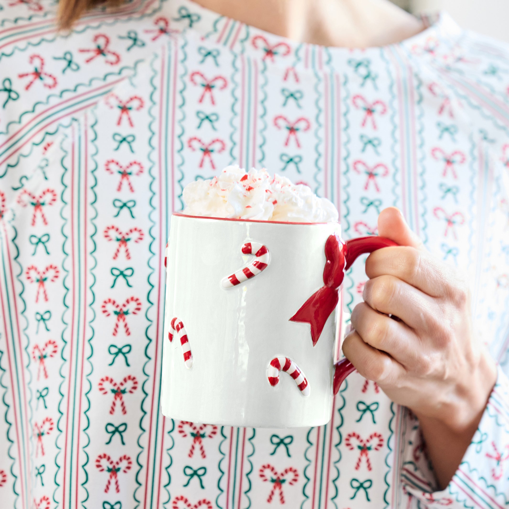 A person wearing a festive holiday sweater with a red, green, and white candy cane and bow pattern, holding a white mug filled with whipped cream and a red handle.