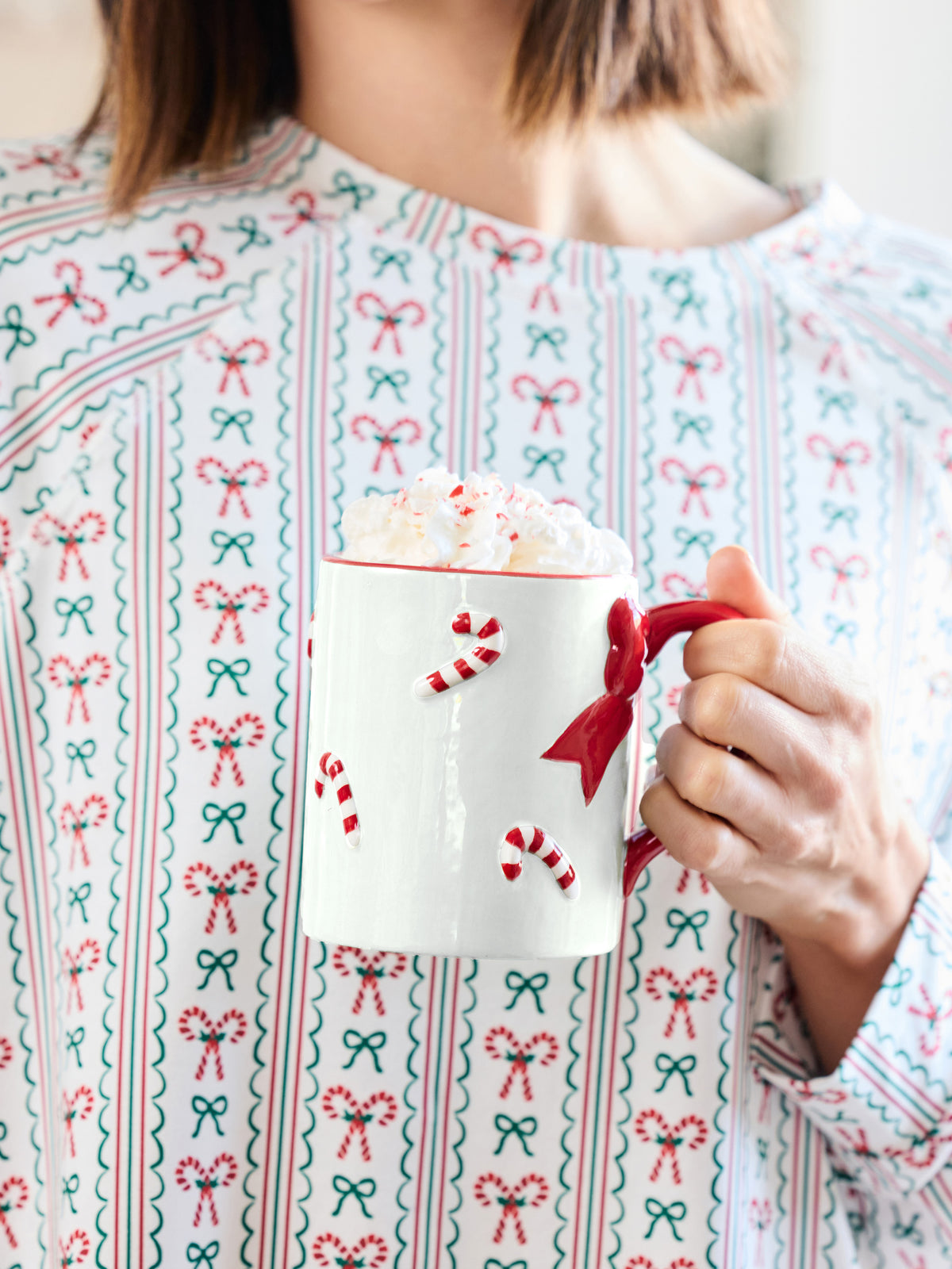A person wearing a festive holiday sweater with a red, green, and white candy cane and bow pattern, holding a white mug filled with whipped cream and a red handle.