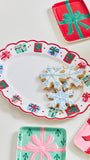 A festive holiday serving platter with a scalloped red border and printed gift box motifs, styled with snowflake-shaped cookies on a white surface. Partially visible matching gift plate designs in red, mint green, and pink surround the platter.