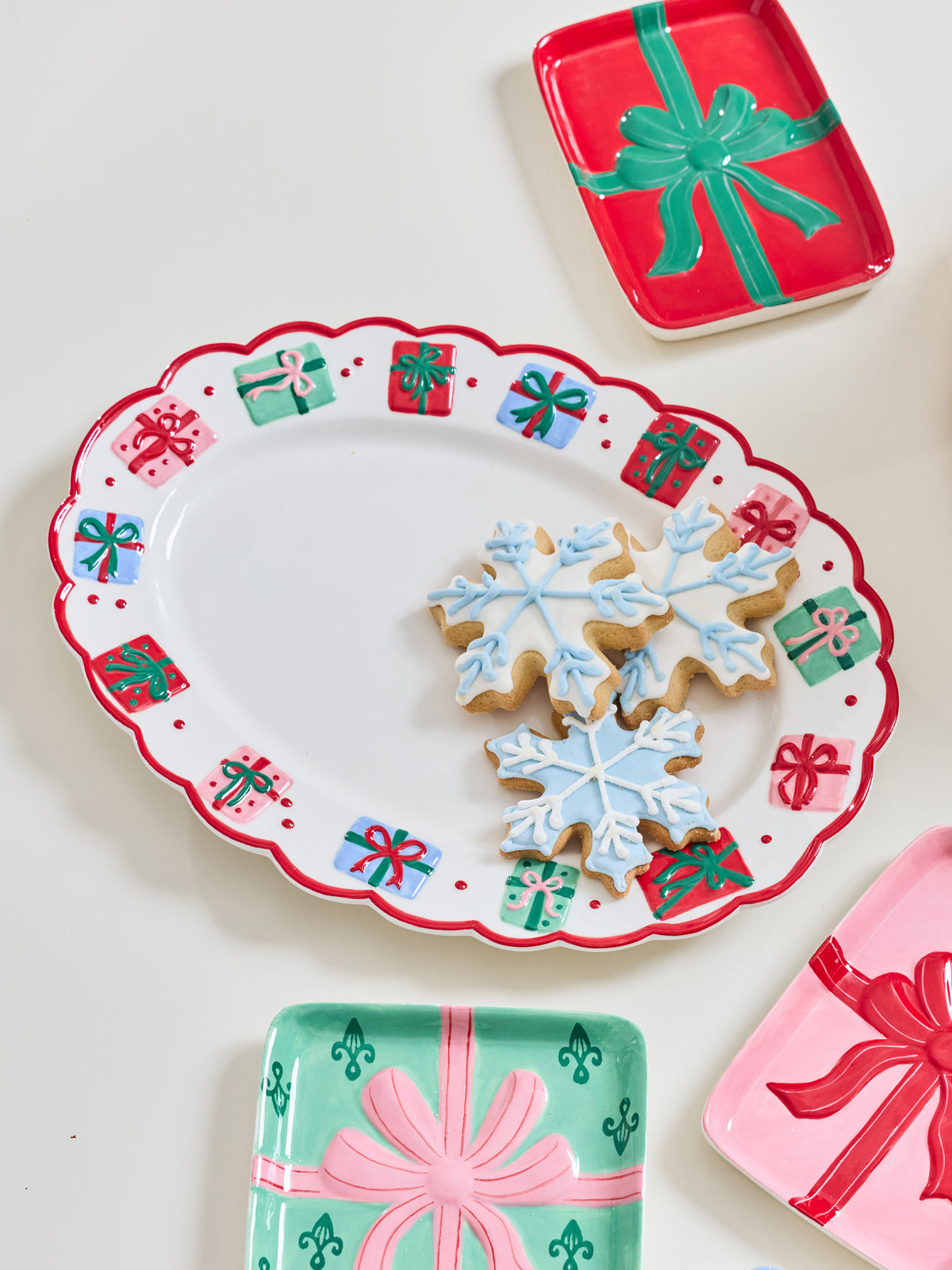 A festive holiday serving platter with a scalloped red border and printed gift box motifs, styled with snowflake-shaped cookies on a white surface. Partially visible matching gift plate designs in red, mint green, and pink surround the platter.