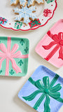 Three decorative square trays shaped like wrapped gift boxes are arranged on a light cream surface. One tray is mint green with a pink bow and small green gift motifs, one is pink with a red bow, and one is light blue with a green bow.