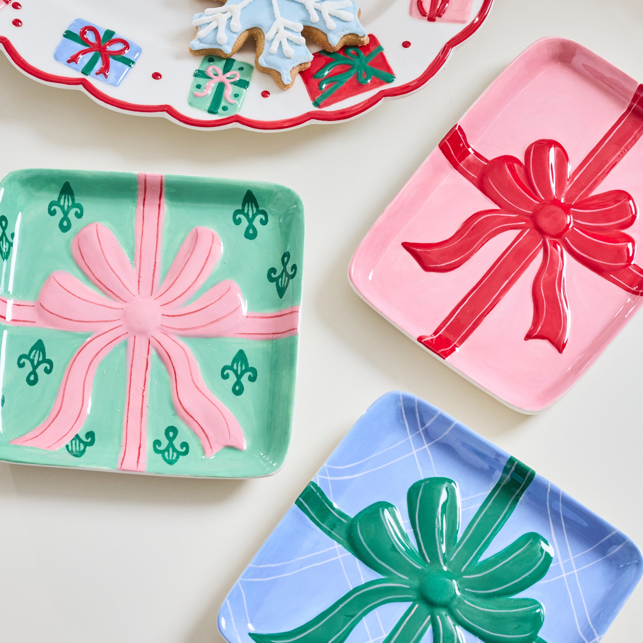 Three decorative square trays shaped like wrapped gift boxes are arranged on a light cream surface. One tray is mint green with a pink bow and small green gift motifs, one is pink with a red bow, and one is light blue with a green bow.