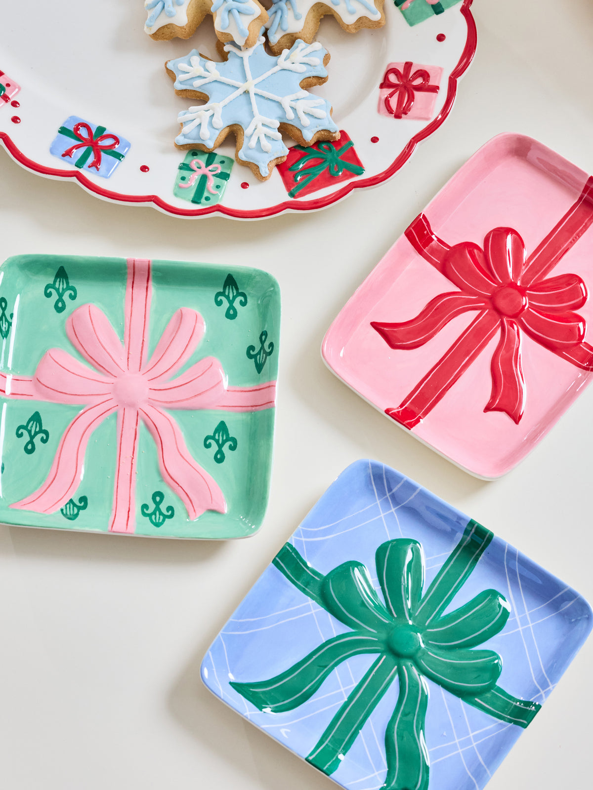 Three decorative square trays shaped like wrapped gift boxes are arranged on a light cream surface. One tray is mint green with a pink bow and small green gift motifs, one is pink with a red bow, and one is light blue with a green bow.
