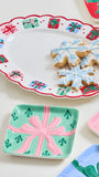 A set of ceramic holiday serving dishes with festive gift-wrap patterns, shown styled with snowflake-shaped cookies on a white background.