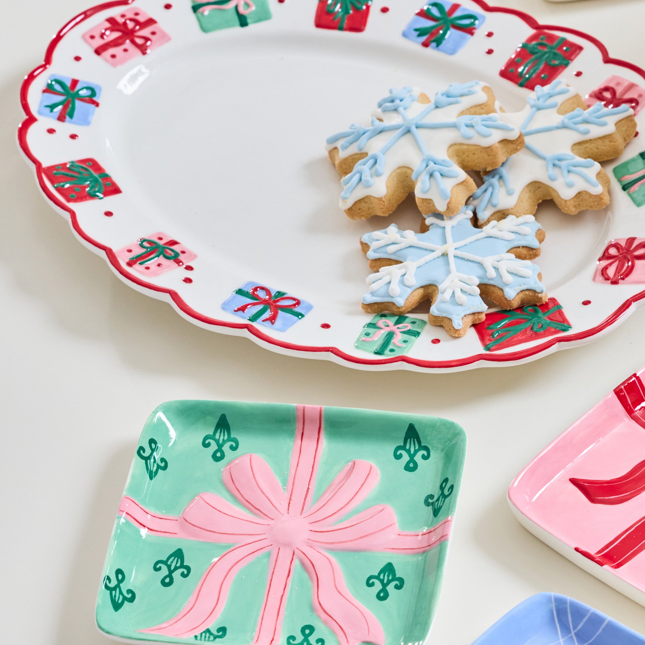 A set of ceramic holiday serving dishes with festive gift-wrap patterns, shown styled with snowflake-shaped cookies on a white background.