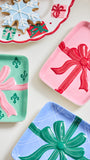 Three decorative holiday serving trays with embossed gift box designs, shown on a light cream background. One tray is mint green with a pink ribbon and green fleur-de-lis motifs, one is pink with a red ribbon and bow, and one is light blue with a green ribbon and bow.
