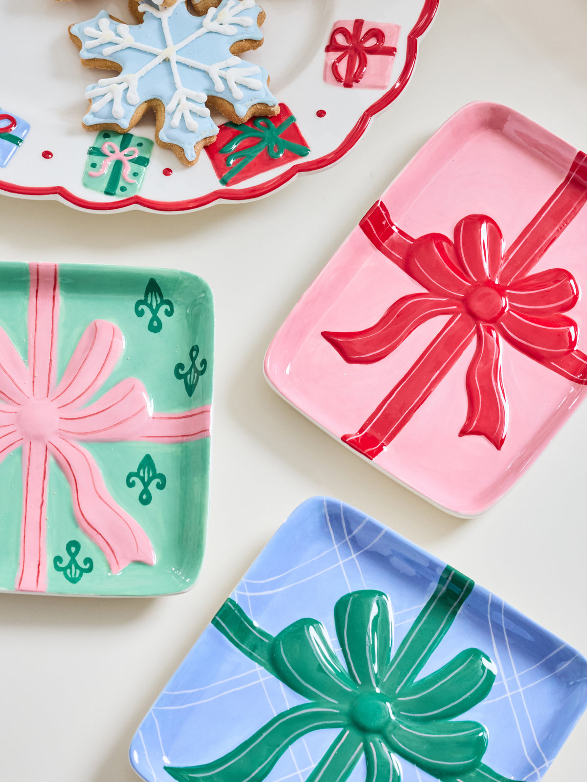 Three decorative holiday serving trays with embossed gift box designs, shown on a light cream background. One tray is mint green with a pink ribbon and green fleur-de-lis motifs, one is pink with a red ribbon and bow, and one is light blue with a green ribbon and bow.