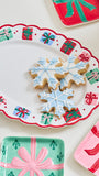 A festive holiday serving platter with a scalloped white rim decorated with red and green gift box motifs, holding three snowflake-shaped iced cookies. Partially visible matching gift-themed plates in red, pink, and mint green surround it on a light cream background.