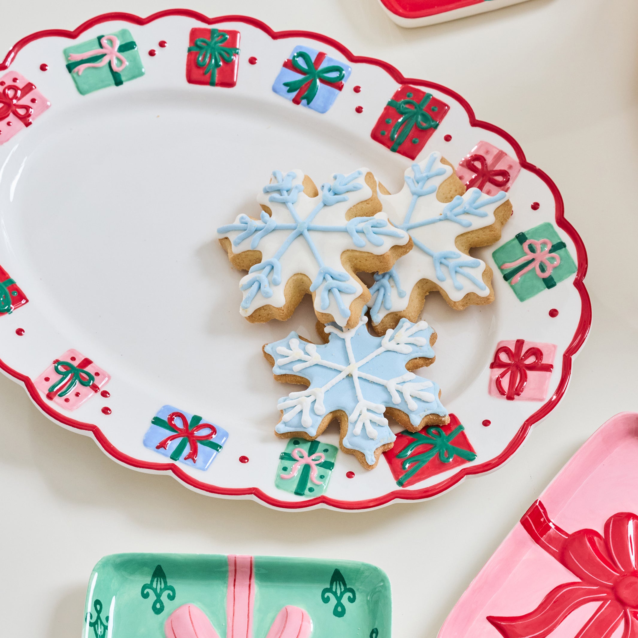 A festive holiday serving platter with a scalloped white rim decorated with red and green gift box motifs, holding three snowflake-shaped iced cookies. Partially visible matching gift-themed plates in red, pink, and mint green surround it on a light cream background.
