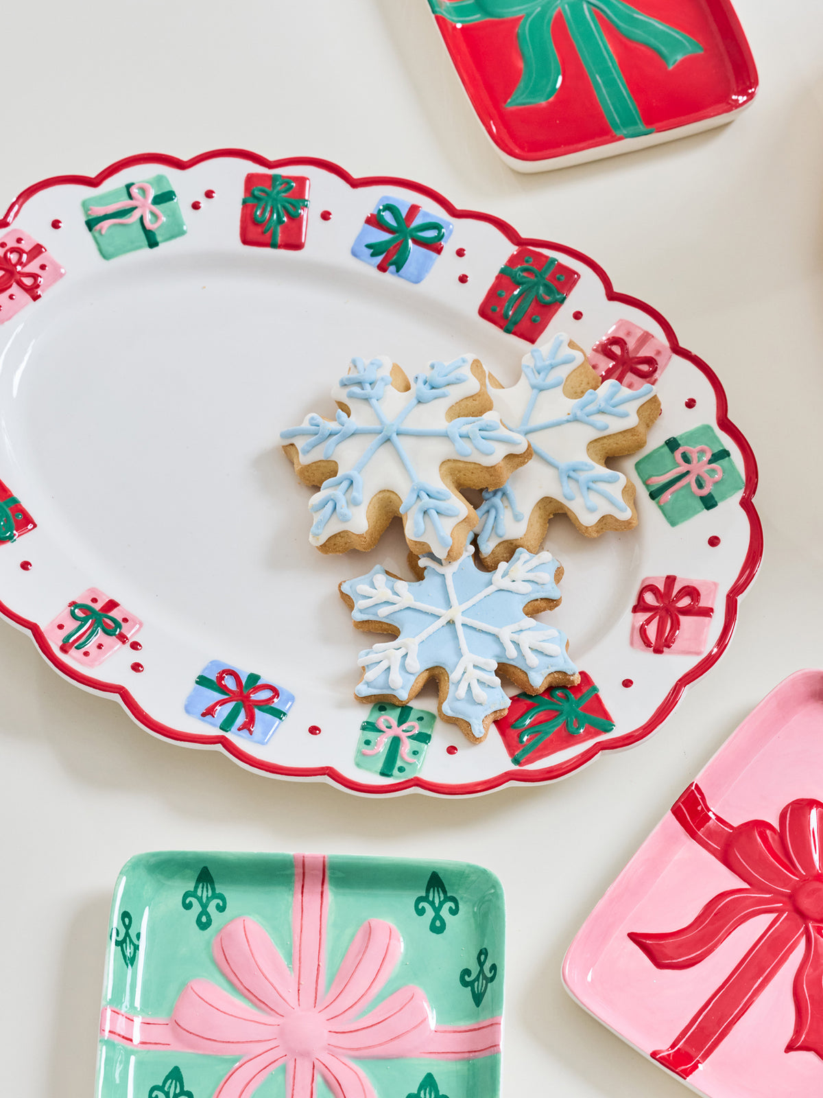 A festive holiday serving platter with a scalloped white rim decorated with red and green gift box motifs, holding three snowflake-shaped iced cookies. Partially visible matching gift-themed plates in red, pink, and mint green surround it on a light cream background.