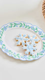 A white ceramic serving platter with a scalloped edge, decorated with a border of blue flowers and green leaves. Three snowflake-shaped sugar cookies with light blue icing are arranged in the center of the platter.