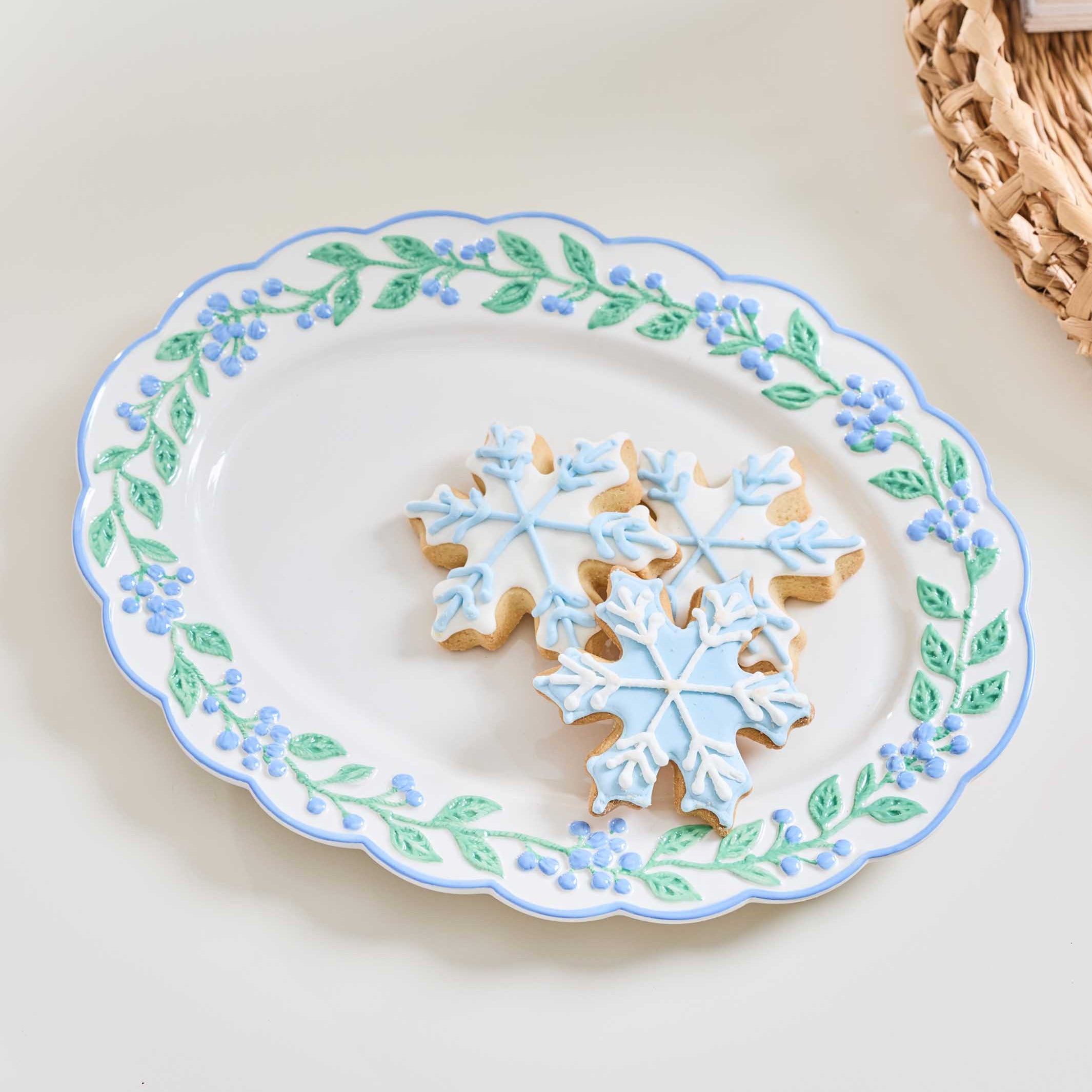 A white ceramic serving platter with a scalloped edge, decorated with a border of blue flowers and green leaves. Three snowflake-shaped sugar cookies with light blue icing are arranged in the center of the platter.