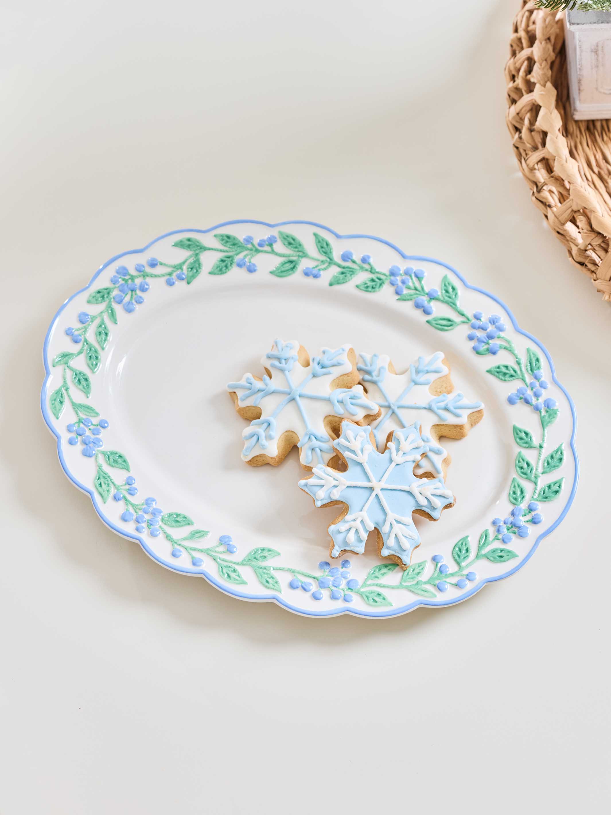 A white ceramic serving platter with a scalloped edge, decorated with a border of blue flowers and green leaves. Three snowflake-shaped sugar cookies with light blue icing are arranged in the center of the platter.