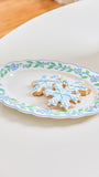 An oval white ceramic plate with a blue and green floral border, holding two snowflake-shaped sugar cookies decorated with light blue icing, set on a white table.