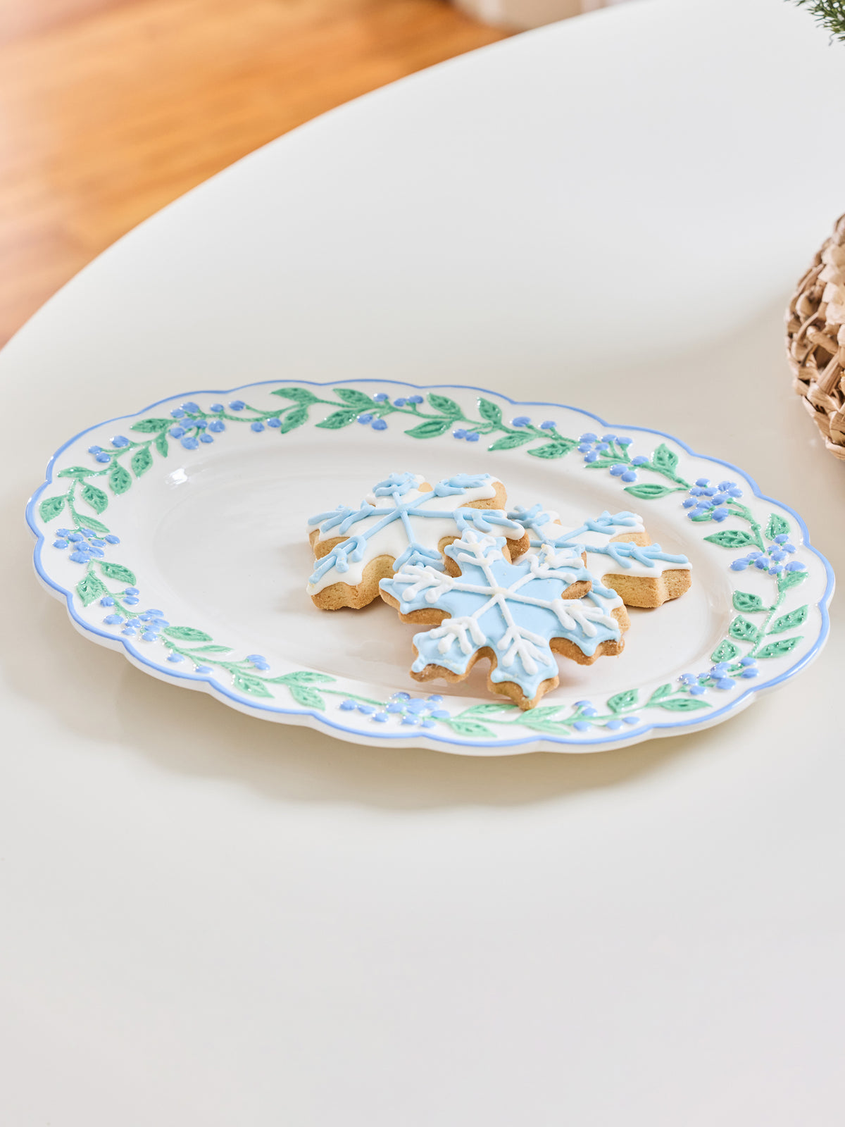 An oval white ceramic plate with a blue and green floral border, holding two snowflake-shaped sugar cookies decorated with light blue icing, set on a white table.