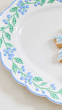 A close-up of a white ceramic plate with a scalloped edge, decorated with a light blue rim and a green leafy vine pattern with small blue berries. A partially decorated gingerbread cookie with blue icing sits on the plate against a warm cream background.