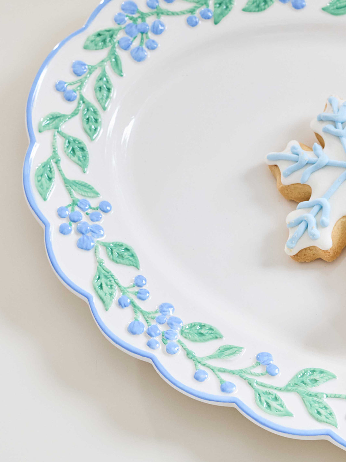 A close-up of a white ceramic plate with a scalloped edge, decorated with a light blue rim and a green leafy vine pattern with small blue berries. A partially decorated gingerbread cookie with blue icing sits on the plate against a warm cream background.