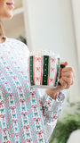 A woman in a festive holiday-themed long-sleeve top holds a white mug decorated with green and red candy cane and bow patterns. The background is a softly blurred indoor setting with warm lighting.