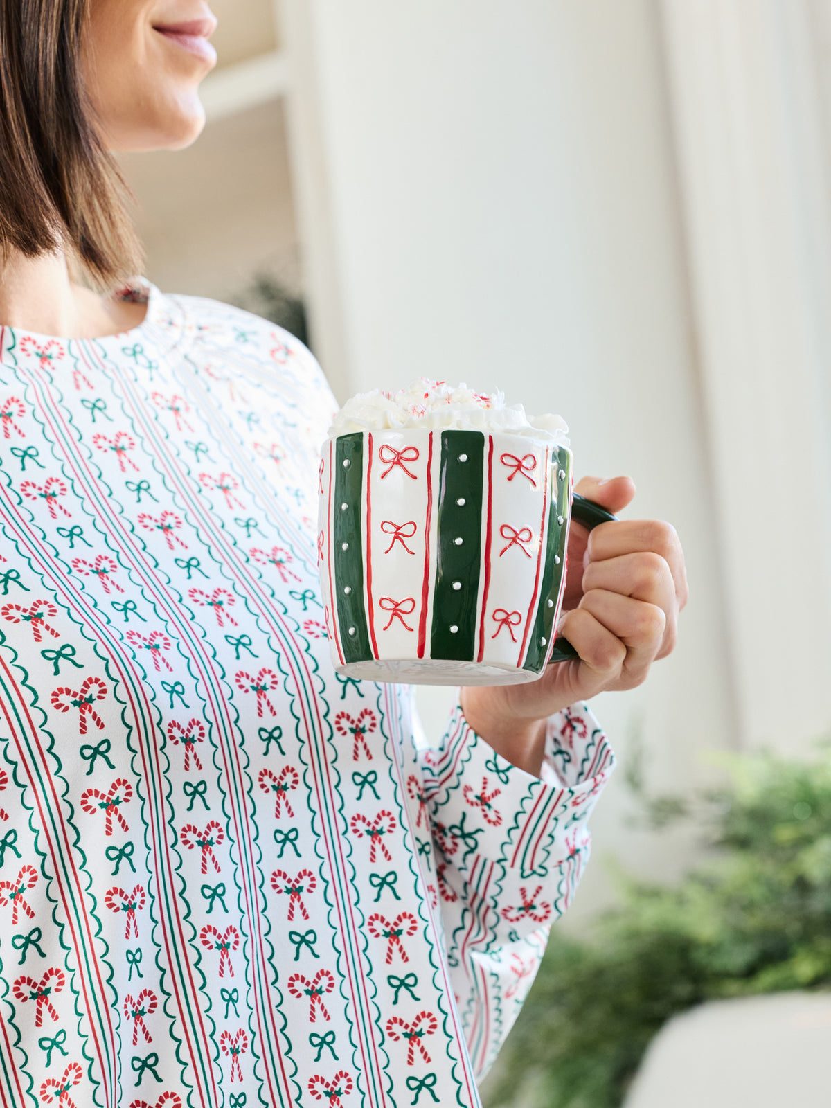 A woman in a festive holiday-themed long-sleeve top holds a white mug decorated with green and red candy cane and bow patterns. The background is a softly blurred indoor setting with warm lighting.