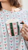 A person in a festive holiday sweater holds a white ceramic mug with green and red candy cane and bow patterns, topped with whipped cream and marshmallows.