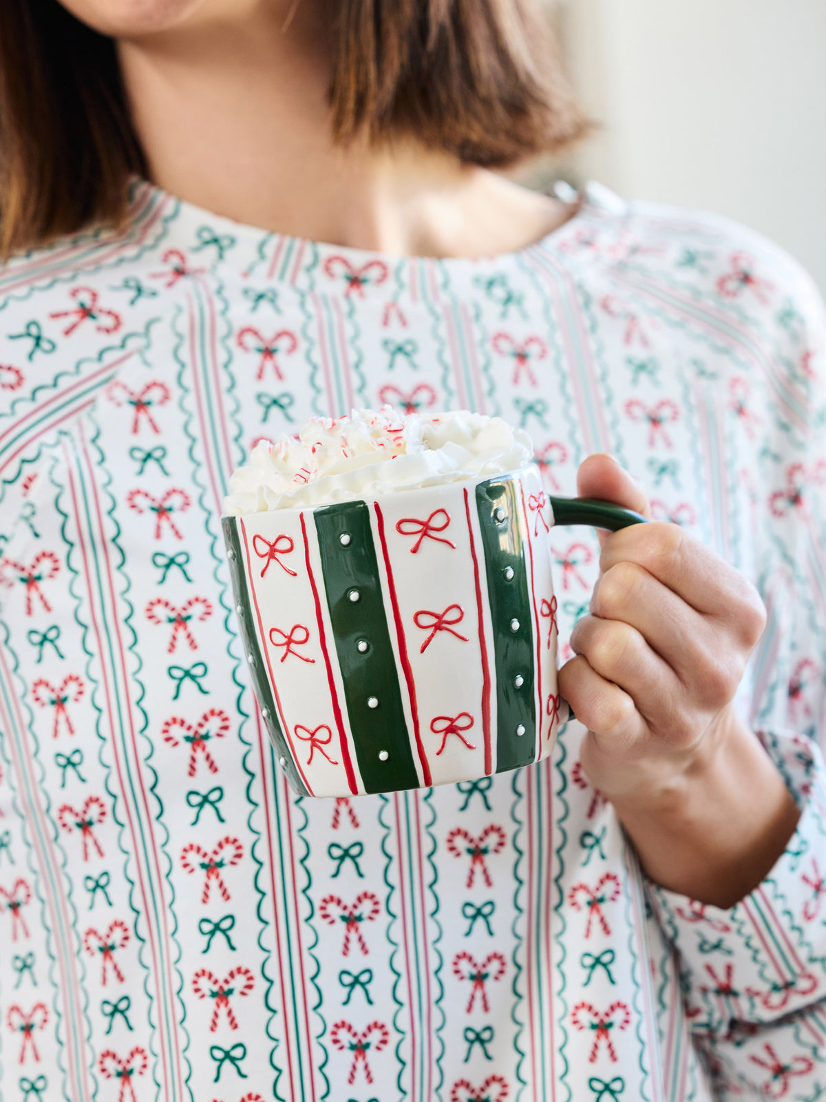 A person in a festive holiday sweater holds a white ceramic mug with green and red candy cane and bow patterns, topped with whipped cream and marshmallows.