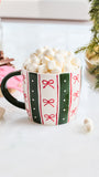 A festive ceramic mug filled with mini marshmallows, decorated with green and red vertical stripes and red bow motifs on a white background. The mug is styled with holiday props including a red and white striped napkin, cinnamon sticks, marshmallows, and evergreen branches.