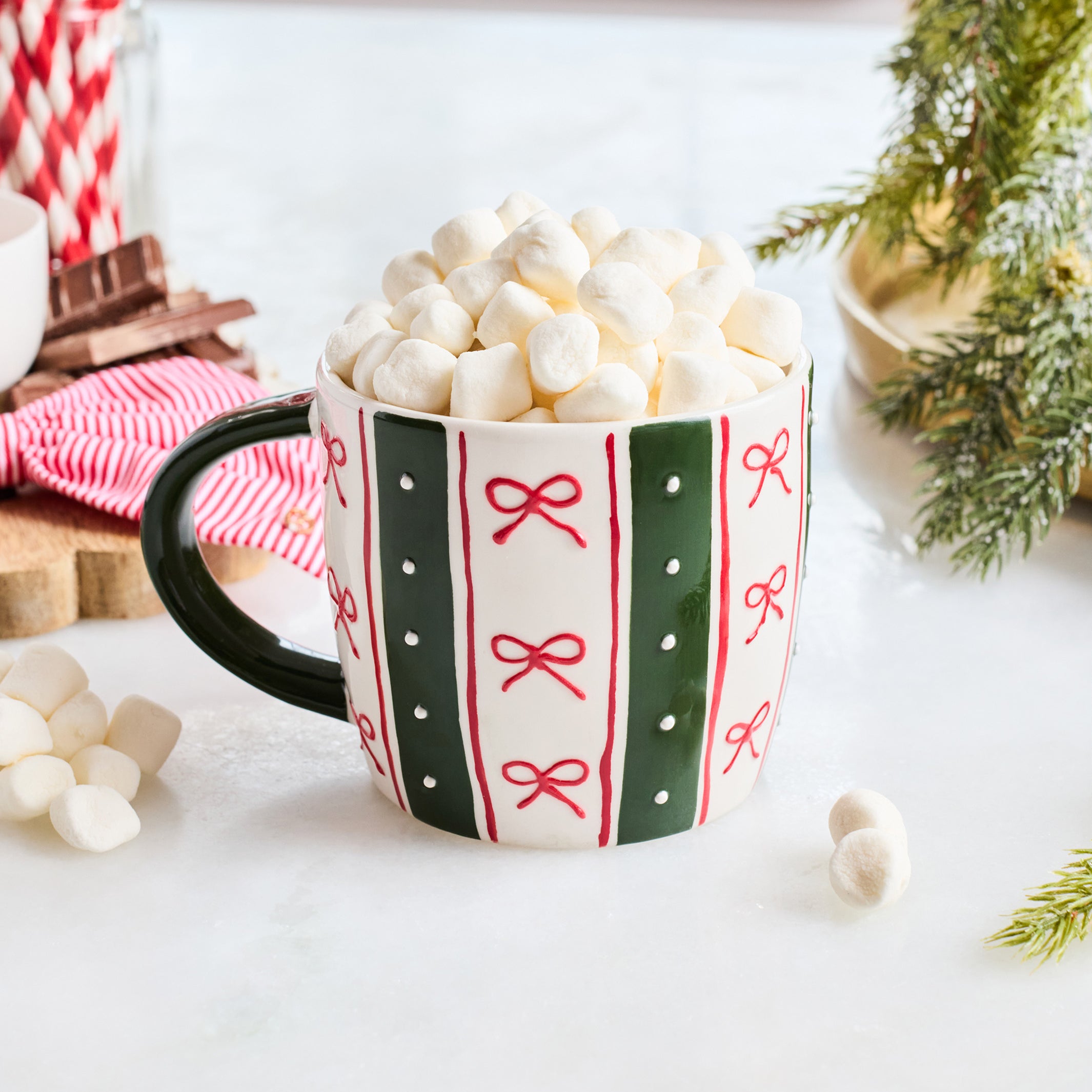 A festive ceramic mug filled with mini marshmallows, decorated with green and red vertical stripes and red bow motifs on a white background. The mug is styled with holiday props including a red and white striped napkin, cinnamon sticks, marshmallows, and evergreen branches.