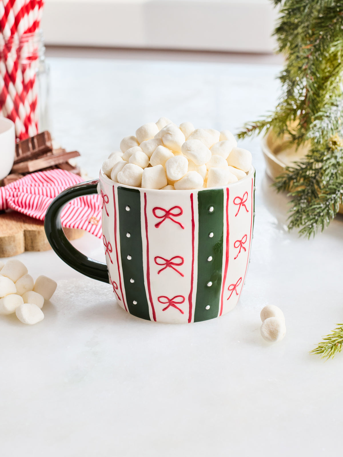 A festive ceramic mug filled with mini marshmallows, decorated with green and red vertical stripes and red bow motifs on a white background. The mug is styled with holiday props including a red and white striped napkin, cinnamon sticks, marshmallows, and evergreen branches.