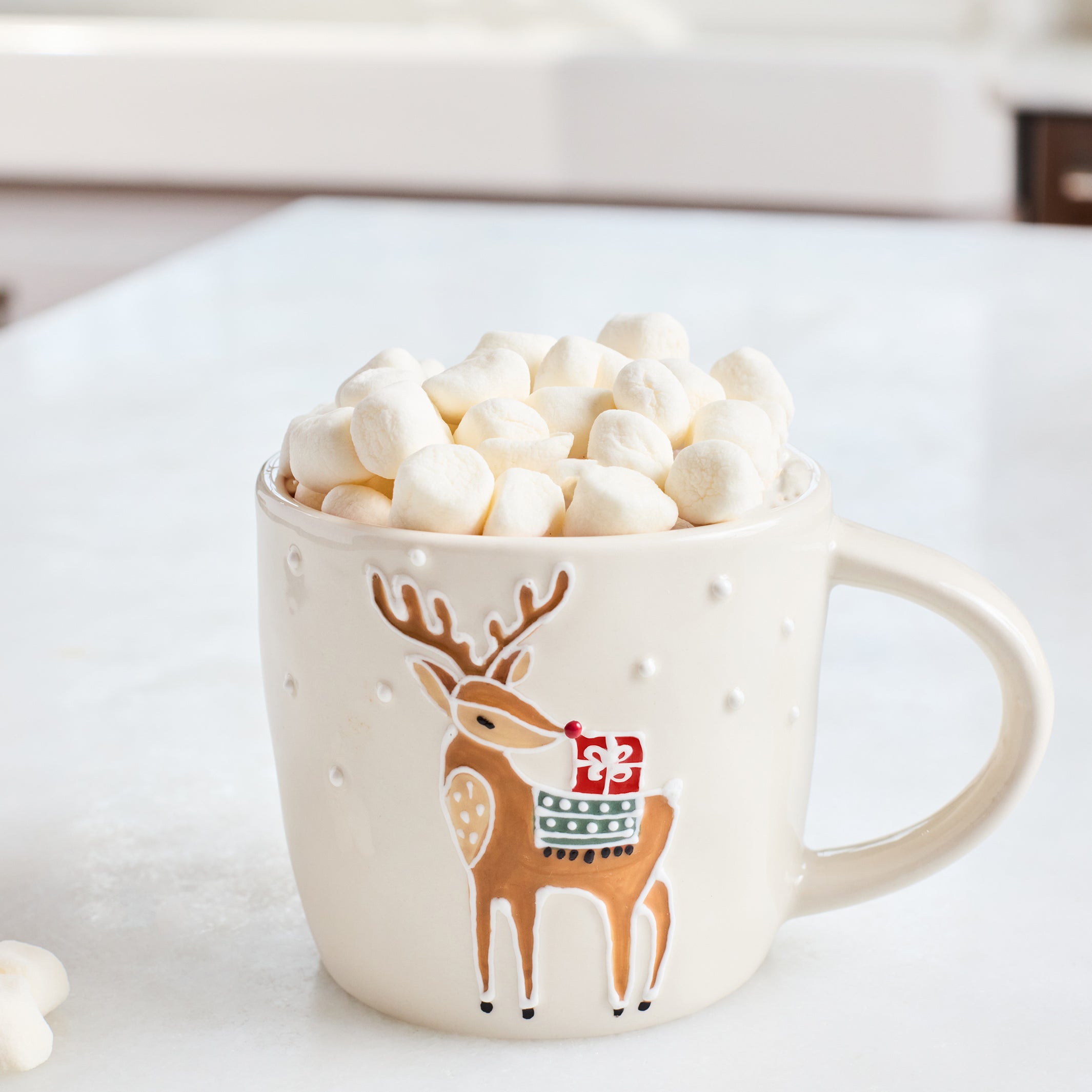 A cream-colored ceramic mug with a brown deer design wearing a red Santa hat, filled with mini marshmallows on a white countertop.