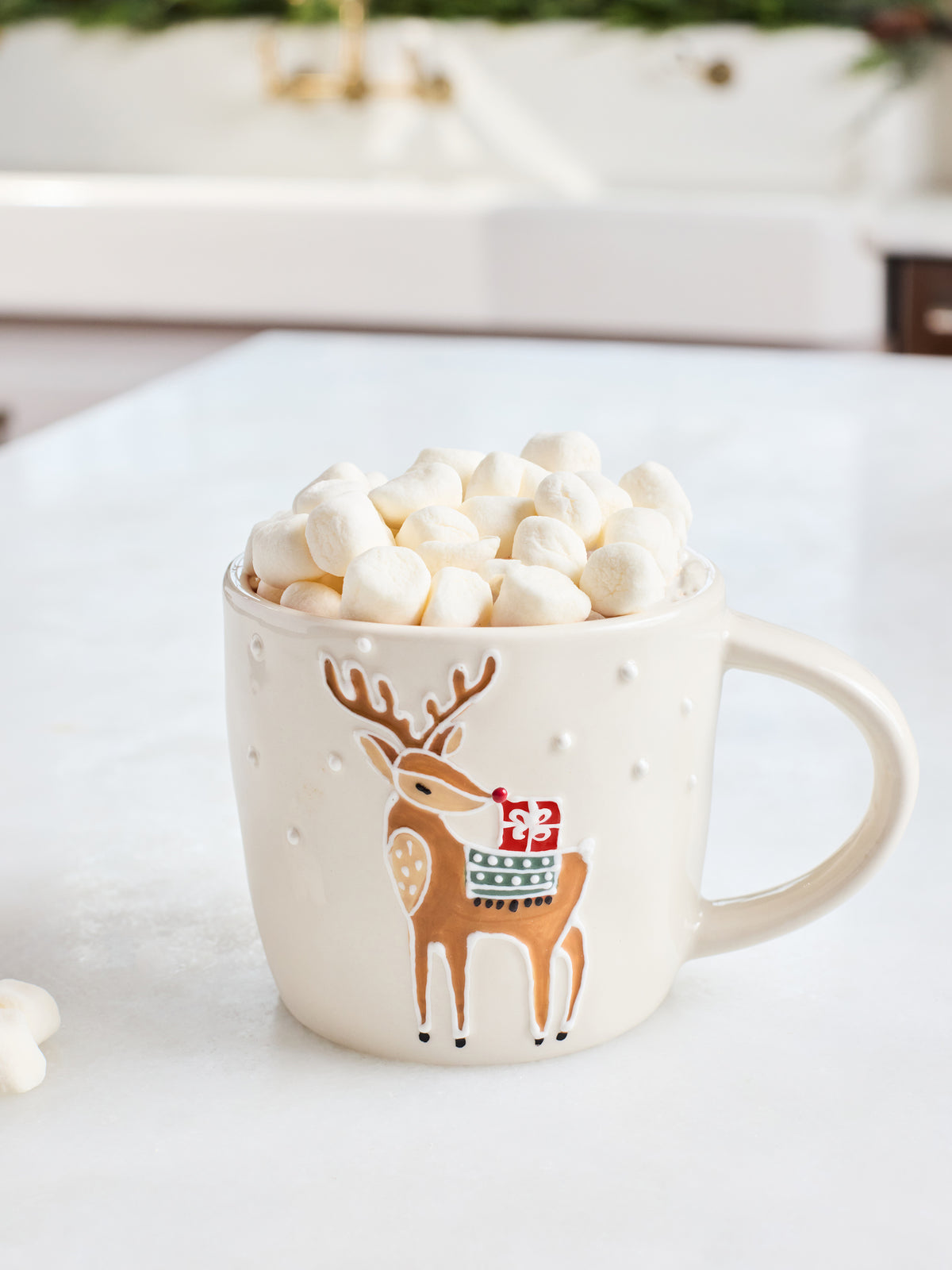 A cream-colored ceramic mug with a brown deer design wearing a red Santa hat, filled with mini marshmallows on a white countertop.
