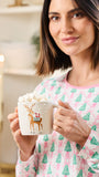 A woman in pink and green pajamas holds a white mug filled with whipped cream and marshmallows. The mug features a gold reindeer carrying a red gift box.