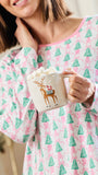 A woman wearing a pink and green holiday-themed long-sleeve pajama top holds a white mug filled with a hot drink and marshmallows. The pajama top features an all-over pattern of green Christmas trees, pink bows, and small red ornaments on a white background.